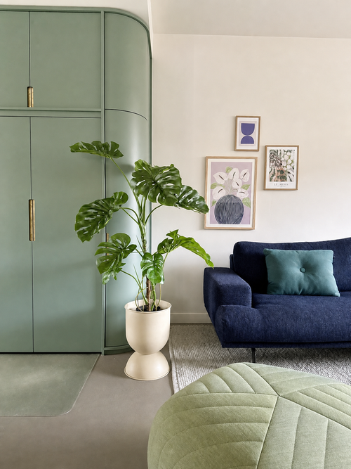 Un soggiorno moderno con un armadio a muro verde, pianta artificiale Monstera 120 cm, un divano blu con un cuscino verde acqua, opere d'arte astratta sul muro e un pouf verde su un tappeto chiaro.
