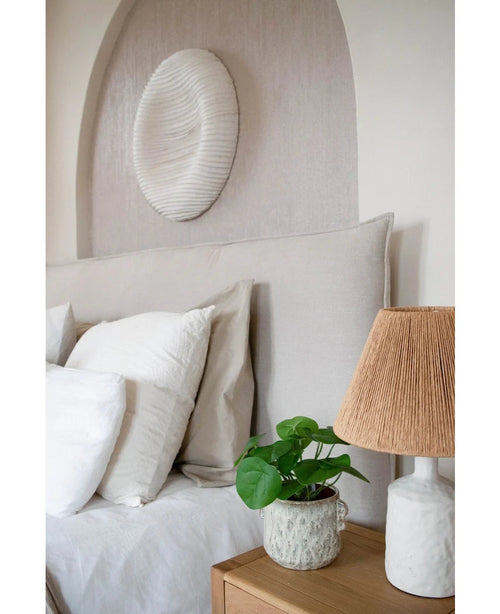 Un accogliente angolo notte con testiera beige, cuscini bianchi e beige, un comodino in legno, una lampada in ceramica con paralume intrecciato, la Pianta Pancake Artificiale 27 cm e decorazioni murali bianche sopra il letto.