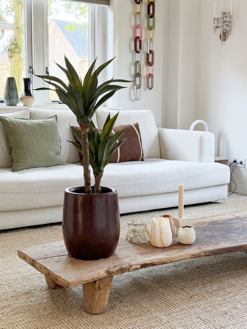 Un soggiorno moderno con un divano bianco, cuscini verdi e marroni, un tavolino in legno decorato con candele e la pianta di Agave artificiale 76 cm, e una decorazione murale a rete. La luce naturale entra dalle finestre dietro il divano.