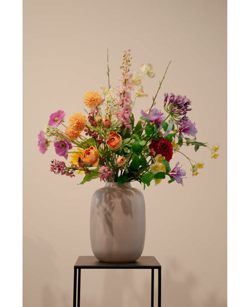 Un vaso beige con ramo di ranuncolo artificiale x5 Real Touch arancione 62 cm ranuncolo arancione artificiale si trova su un supporto in metallo nero su uno sfondo chiaro.