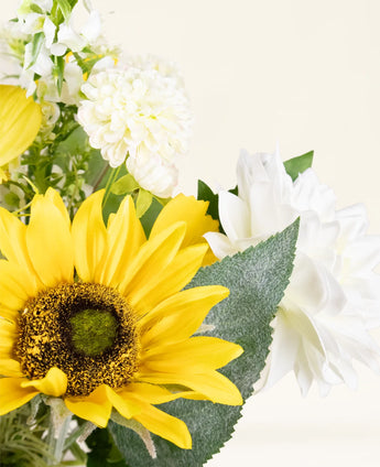 Een close-up van het Zijden kunstbloemen boeket Sunny Whisper met een heldergele zonnebloem, witte dahlia's en groene bladeren tegen een lichte, neutrale achtergrond.