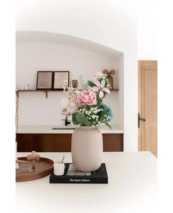 De Vaas Artic Beige Ø21 x H29 cm, gevuld met roze en blauwe kunstbloemen, staat op een zwart boek op een wit aanrecht. Zachte verlichting accentueert het minimalistische tafereel met decorplanken en een houten deur op de achtergrond.