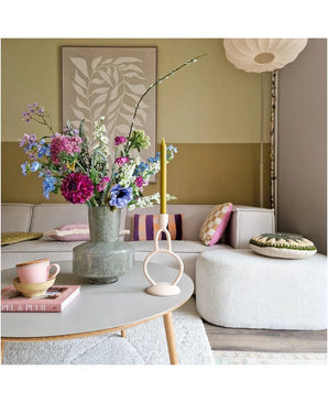 Een gezellige woonkamer met een lichtgrijze bank, kleurrijke kussens en een salontafel met daarop Zijden kunstbloemen boeket Purple Rain-een display van prachtig Nederlands vakmanschap-een kaars en een roze kopje. Een bladerrijk kunstwerk siert de muur.