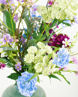 Het zijden kunstbloemen boeket Purple Rain met lichtgroene Queen Anne's lace, blauwe anjers, paarse en roze bloemen in een groene vaas toont Nederlands vakmanschap tegen een witte achtergrond.