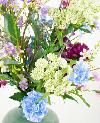 Het zijden kunstbloemen boeket Purple Rain met lichtgroene Queen Anne's lace, blauwe anjers, paarse en roze bloemen in een groene vaas toont Nederlands vakmanschap tegen een witte achtergrond.