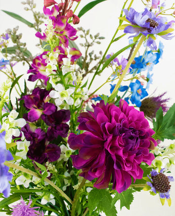 Het Zijden kunstbloemen boeket Purple Rain heeft een grote magenta dahlia, witte en paarse bloemen, blauwe bloesems en groene bladeren, die het Nederlands vakmanschap benadrukken tegen een witte achtergrond.