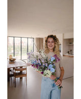 Een vrouw met krullend blond haar en een beige topje glimlacht in een lichte, moderne eetkamer, terwijl ze het Zijden kunstbloemen boeket Purple Rain vasthoudt - een prachtige showcase van Nederlands vakmanschap en levendige kunstbloemen.