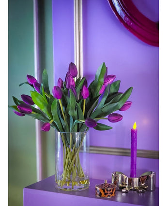 In het Zijden kunstboeket Tulpen in vaas Paars staan levendige paarse tulpen met groene bladeren in een vaas van helder glas, uitgestald op een paars vlak naast een brandende kaars en een decoratieve bak, tegen een lavendelblauwe achtergrond met metallic accenten.