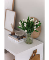 Het Zijden kunstboeket Tulpen in vaas Wit staat op een witte tafel naast gestapelde boeken, een schelpvormig decorstuk en ingelijste kunst tegen een lichtgekleurde muur.