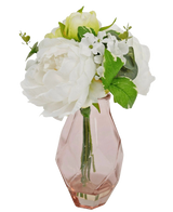 Het Kunstboeket in glas 25 cm heeft een klein boeket van witte bloemen met groene bladeren, gerangschikt in een facetgeslepen, lichtroze glazen vaas.