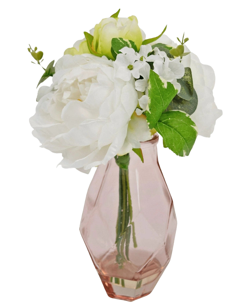 Il Bouquet Artificiale in Vetro 25 cm presenta un piccolo bouquet di fiori bianchi con foglie verdi, disposti in un vaso di vetro sfaccettato di colore rosa chiaro.