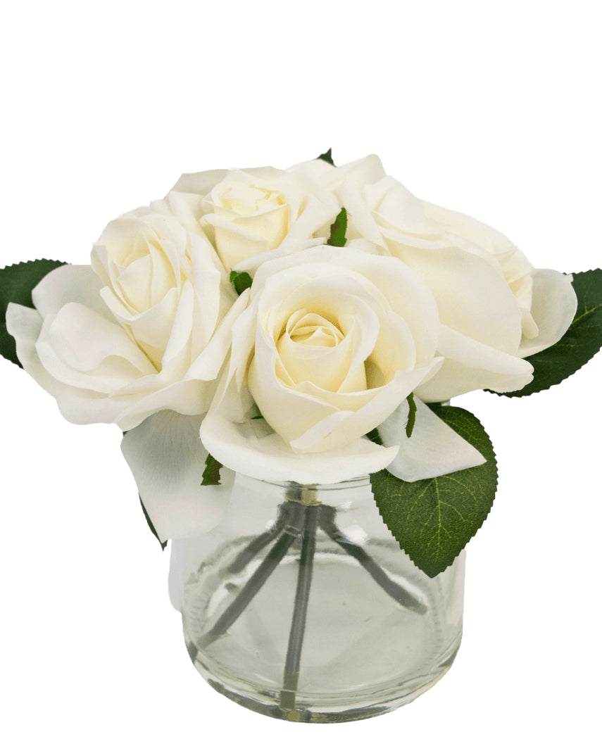 Il bouquet di rose artificiali Real Touch White da 18 cm è un piccolo vaso in vetro trasparente con rose bianche realistiche e foglie verdi, su un semplice sfondo bianco.